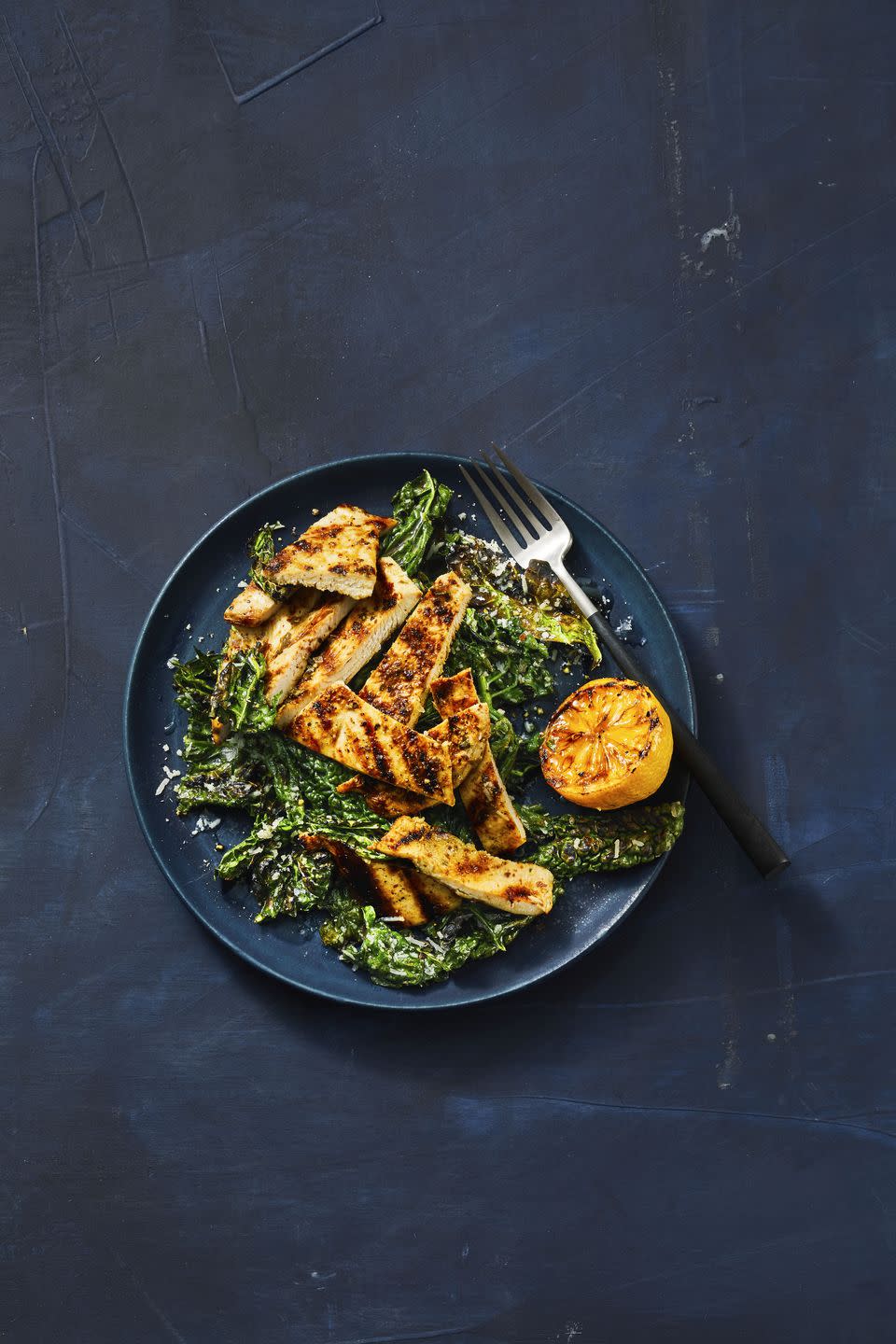 grilled lemony chicken and kale on a dark blue plate with a grilled lemon served on the side