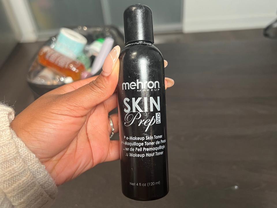 The writer holds a black bottle with white "Mehron skin prep" text