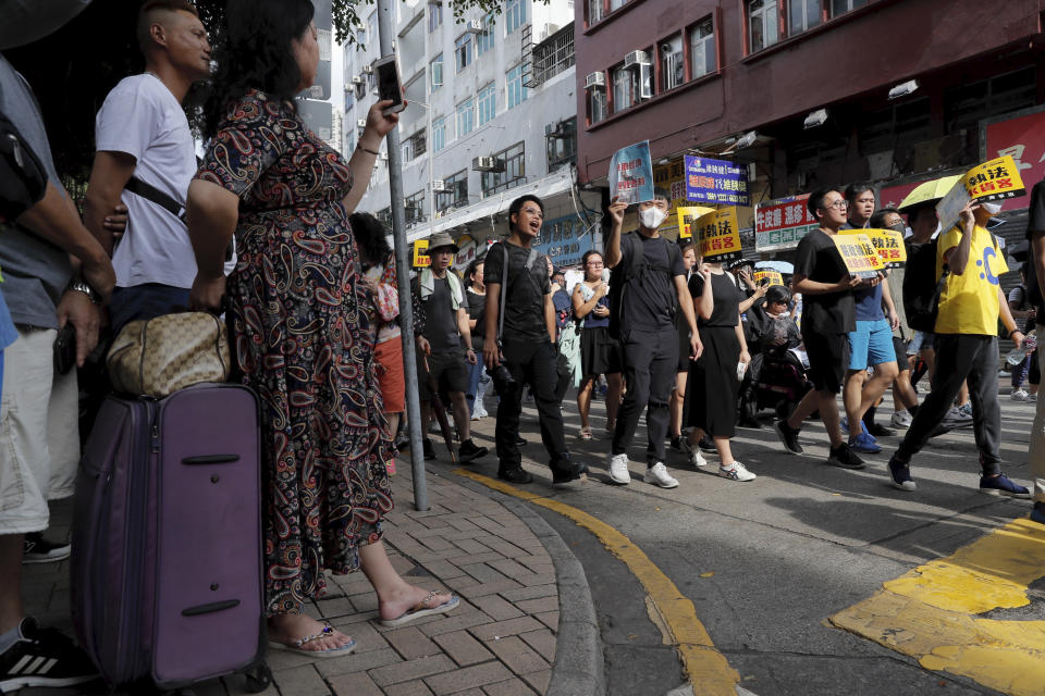 Tourists watch as protesters march in an area popular with Chinese tourists for its pharmacies and cosmetic shops, in Hong Kong, Saturday, July 13, 2019. Several thousand people marched in Hong Kong on Saturday against traders from mainland China in what is fast becoming a summer of unrest in the semi-autonomous Chinese territory. (AP Photo/Kin Cheung)