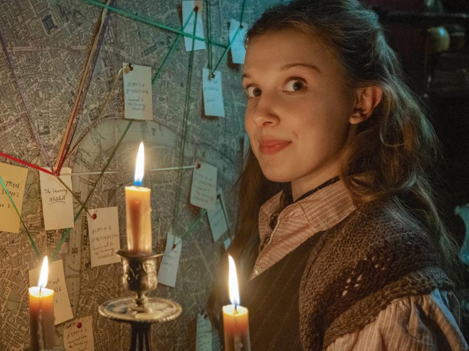 millie bobby brown as enola holmes, standing in front of a map with post-it notes and strings connecting them, illuminating the picture with a candelabra