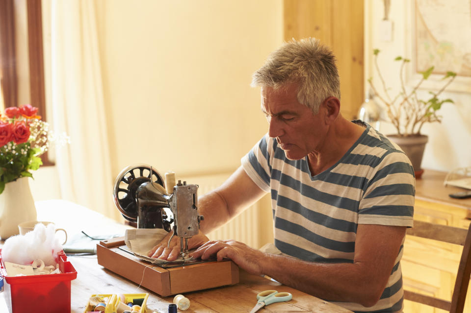 Older man in a striped shirt using a vintage sewing machine at a wooden table with craft supplies around