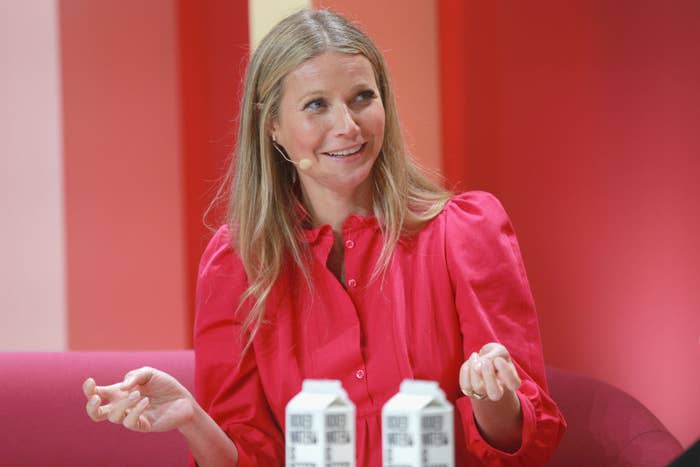 Gwyneth Paltrow in a pink button down in a pink room on a pink couch smiling with a microphone headset