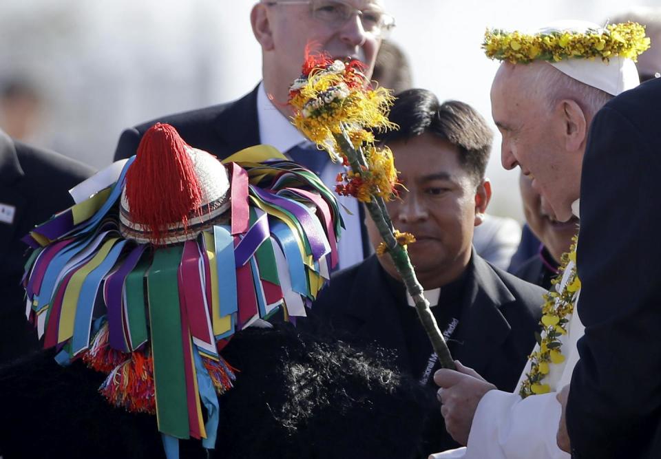 An older man in white wears a crown of yellow flowers, standing amid other men, and near a hat covered in brightly colored ribbons.