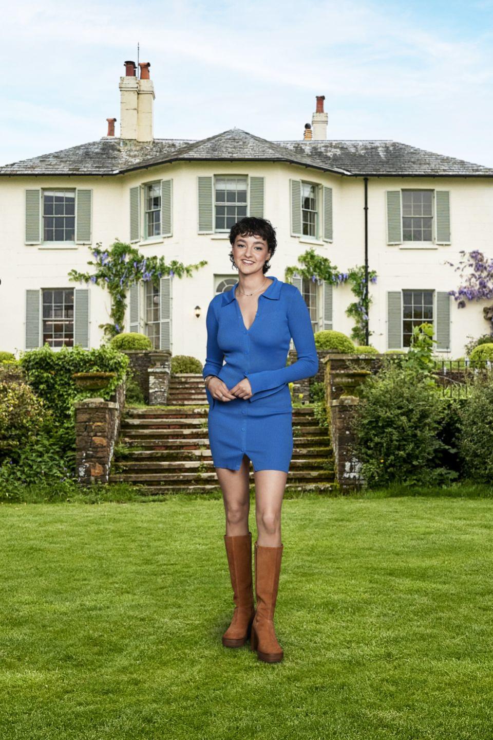 jess from my mum, your dad wearing a blue dress and brown knee high boots, smiling and standing in front of a country estate