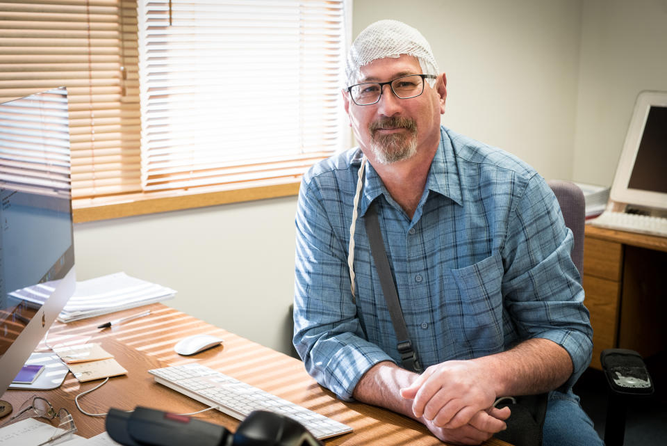 Man sitting at desk wearing NovoCure's Optune, which appears like netting on his head and includes an over-the-shoulder attachment to the head device.