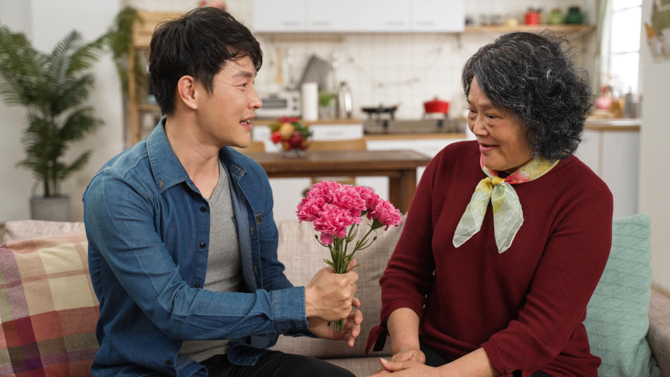 cheerful asian adult son surprising mother with carnation bouquet on Mother’s Day. happy senior woman embracing the man with smile on sofa at home