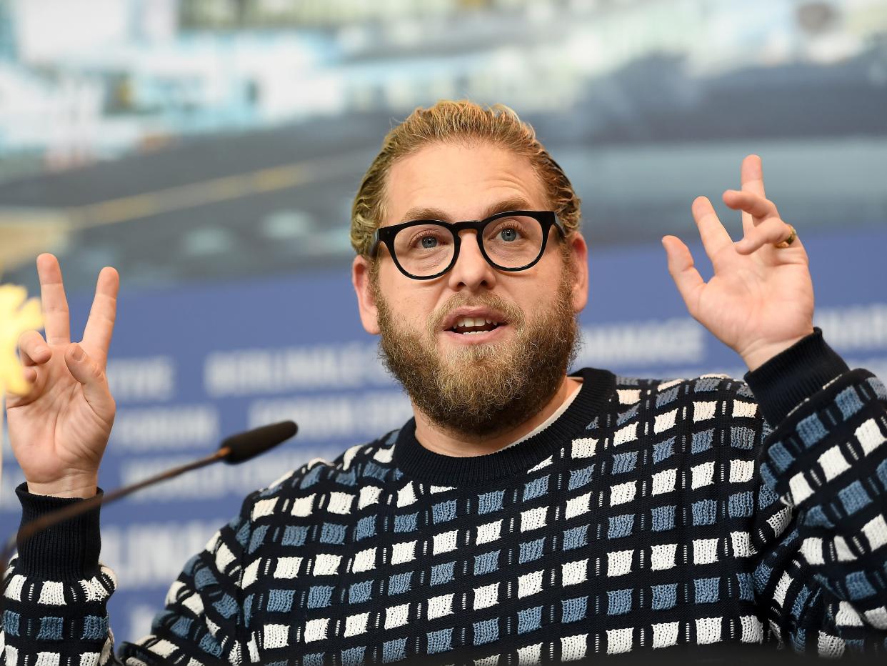 Jonah Hill speaking into a microphone while wearing a black, blue, and white patterned jumper and a pair of glasses.