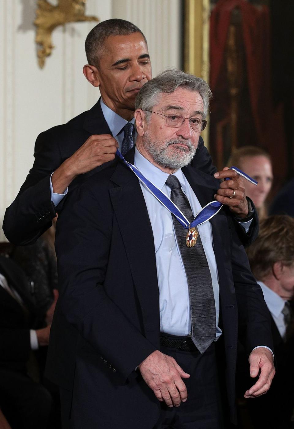 barack obama places a medal around robert de niros neck while both men stand inside a room, they both wear suits with blue collared shirts