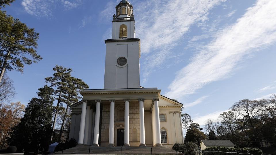 A neoclassical building with a tall clock tower and columns, surrounded by trees and a clear sky