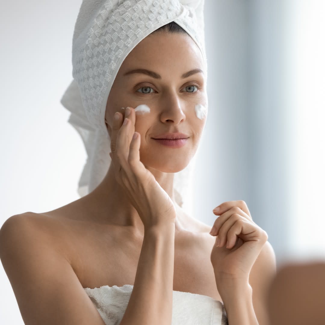  woman applying skincare in her bathroom with a towel on her head 