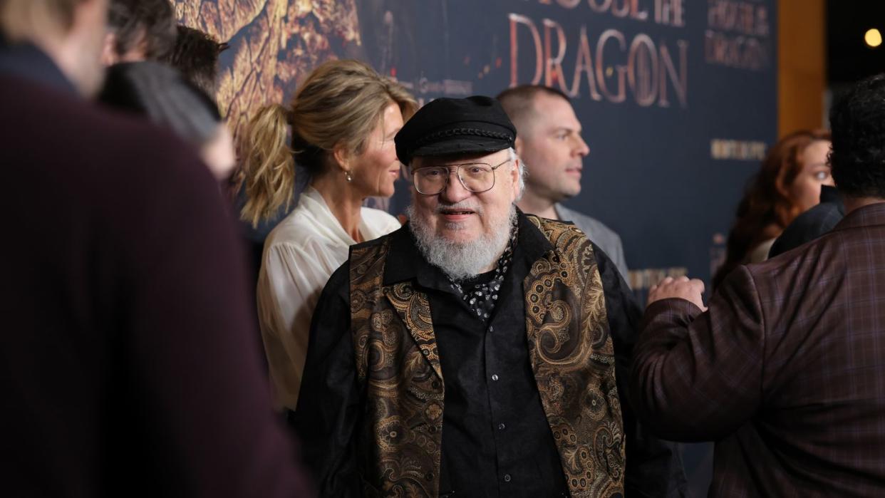 george rr martin smiles as he stands among several over people in front of a backdrop for house of the dragon, martin wears a black hat and button up shirt with a paisley vest over top and his large wire rimmed glasses