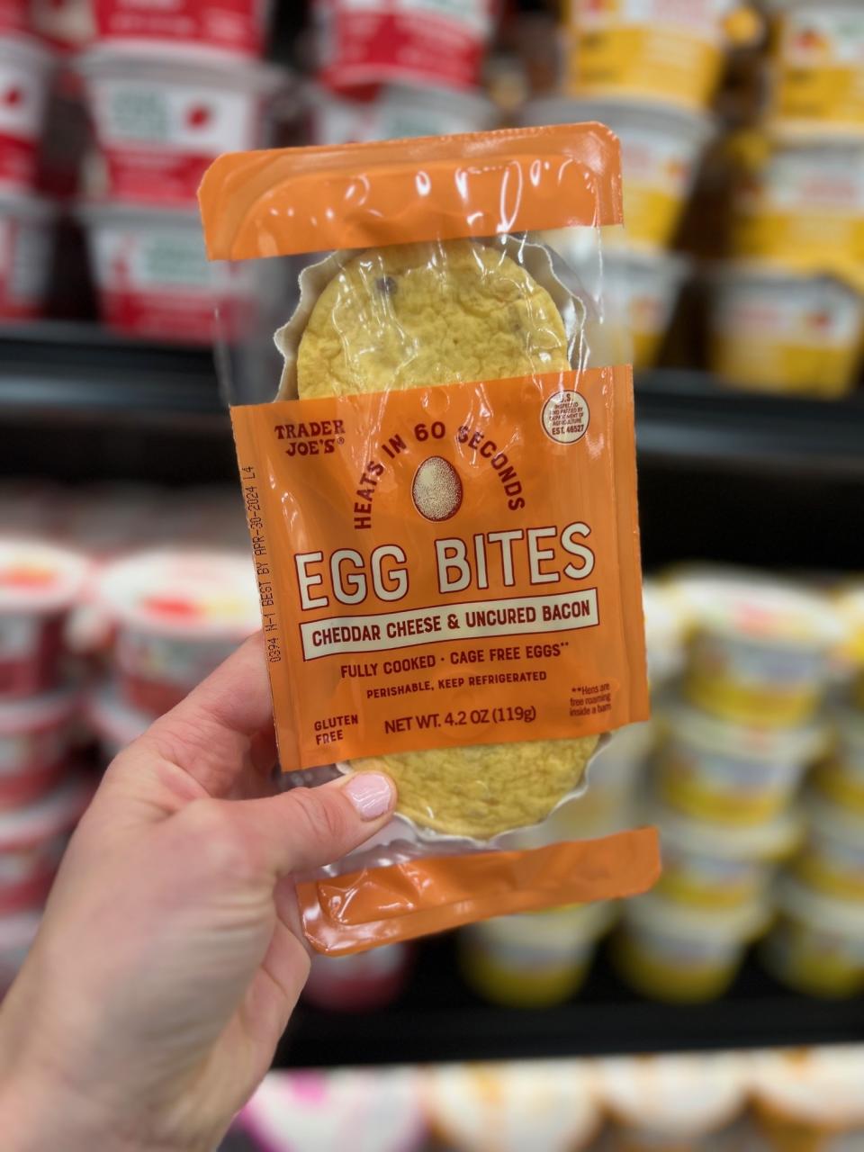 Hand holding a package of Trader Joe's Egg Bites with Cheddar Cheese & Uncured Bacon against store shelves