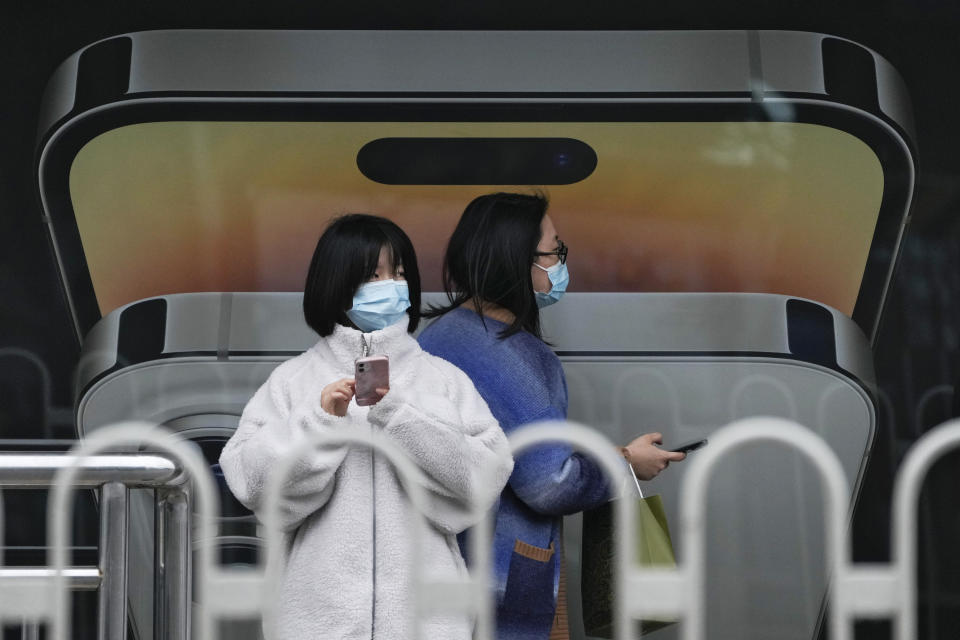 A woman wearing a face mask walks by another using an iPhone as they waiting for their buses at a bus stand displaying an iPhone advertisement in Beijing on Oct. 30, 2022. Access to an industrial zone in the central Chinese city of Zhengzhou was suspended Wednesday after the city reported 64 coronavirus cases and workers who assemble Apple Inc. iPhones left their factory in the zone following outbreaks. (AP Photo/Andy Wong)