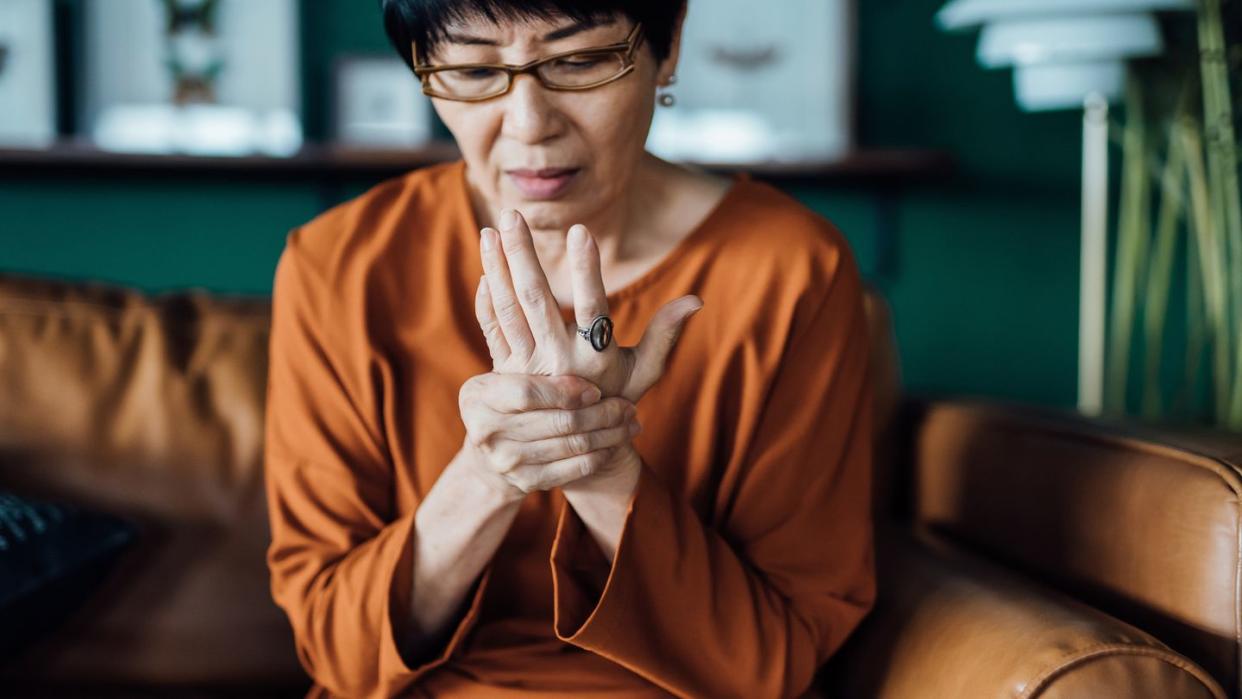 woman rubbing her hands in discomfort, suffering from arthritis in her hand while sitting on sofa at home elderly and health issues concept