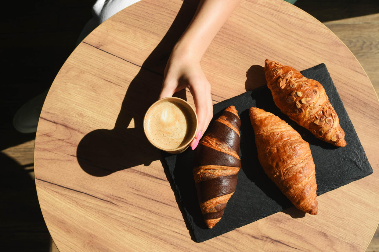 Three croissants  on a small wooden tabletop and a hand holding a cup of butter.