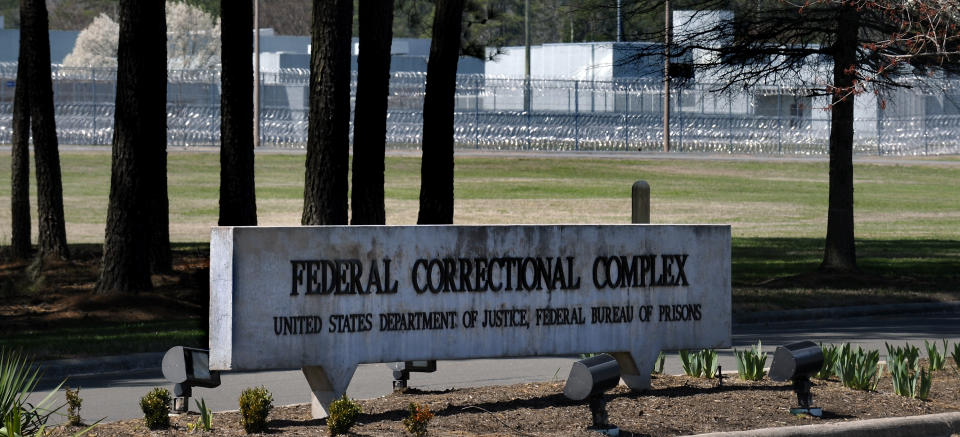 A sign saying Federal Correctional Complex, United States Department of Justice, Federal Bureau of Prisons, stands in front of trees, with a fence protected by voluminous coils of barbed wire in the background.