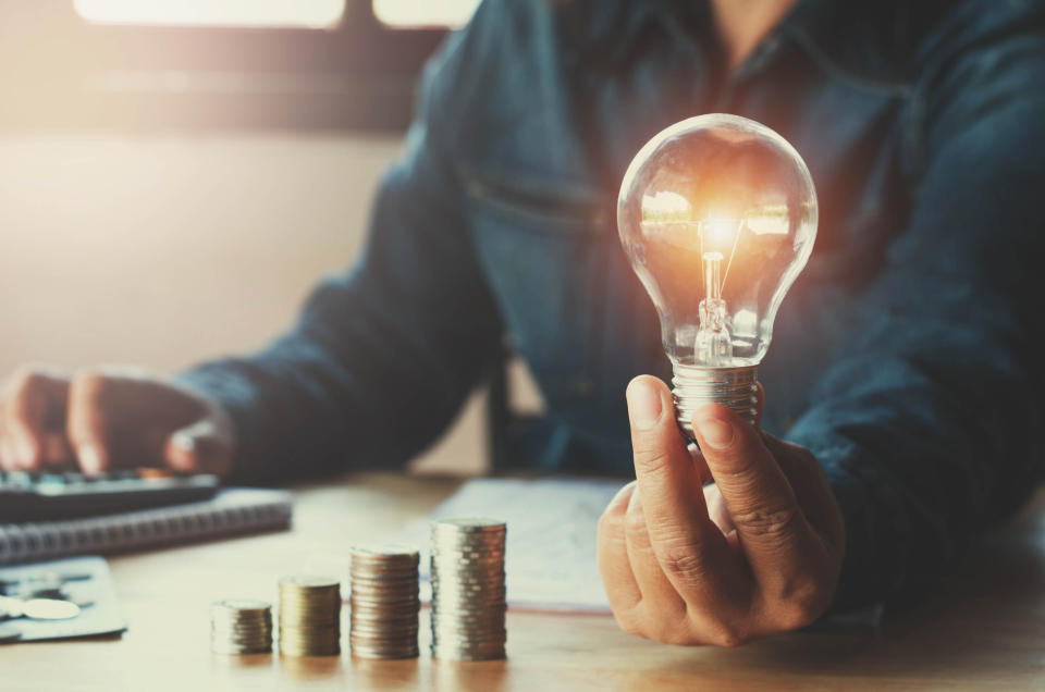 A man sitting at a table holding a light bulb in one hand, with the other hand on a calculator and an ascending stack of coins laying in front of him on the table.