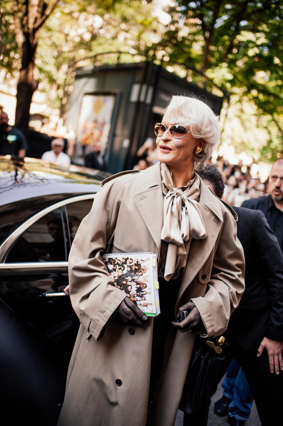 Alexis Stone with short hair and sunglasses, dressed stylishly in a beige trench coat and holding a magazine, stands next to a car in a busy street