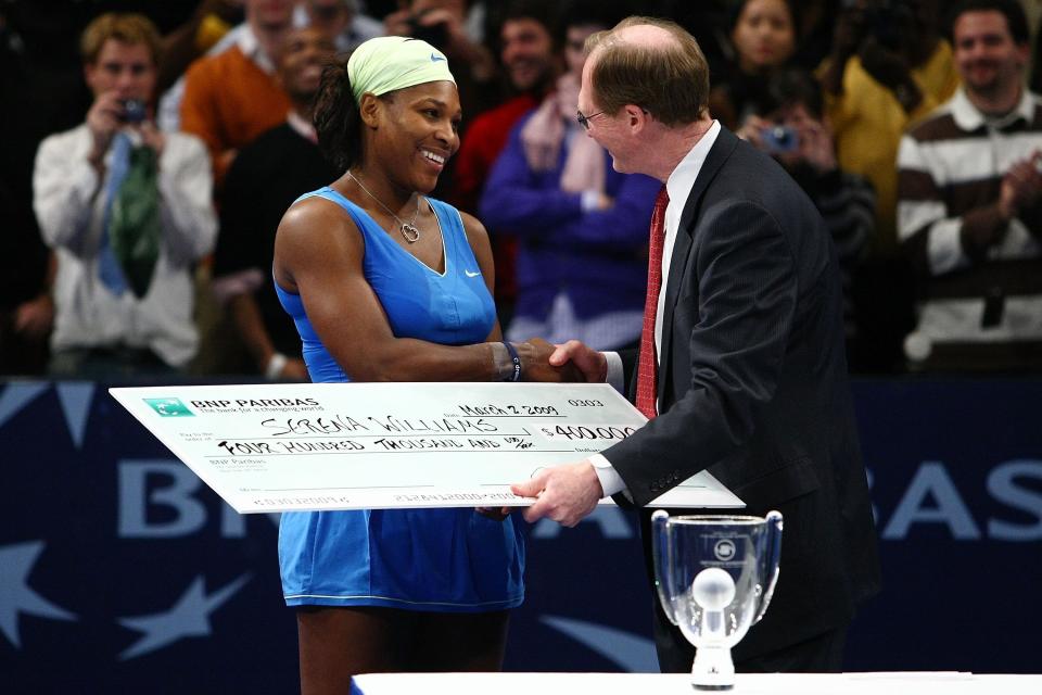 Serena Williams in athletic attire shakes hands with a man in a suit while holding a large check at a tennis event