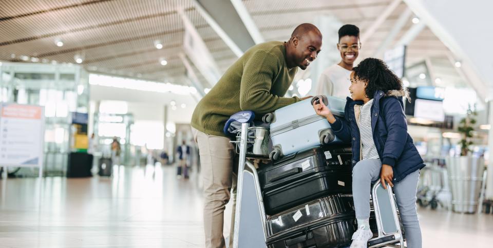 A family at the airport with luggage. The little girl is riding on the stacked luggage cart