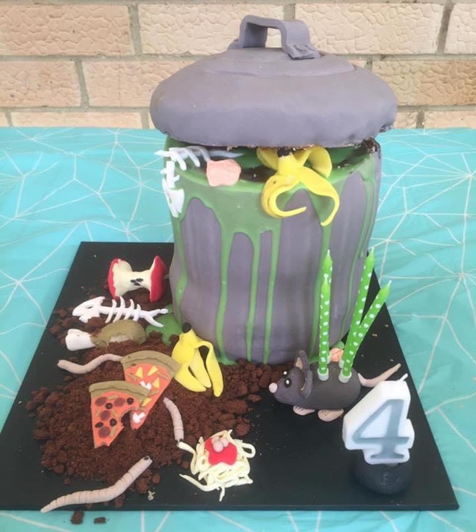 A garbage can birthday cake showing a grey can with overflowing contents including fish bones, pizza, worms and a mouse with four birthday candles in its back.