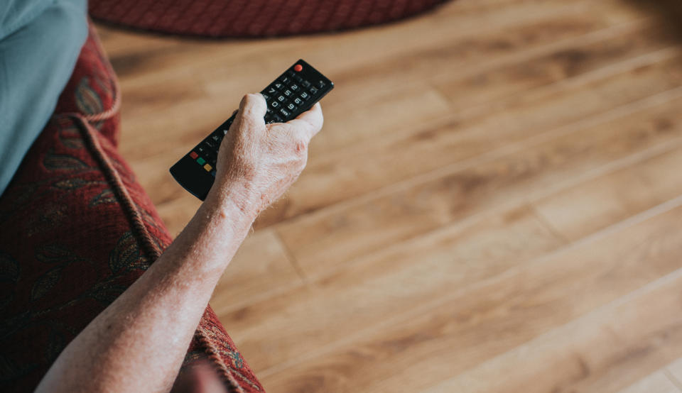 A person holds a TV remote while sitting on a couch. The focus is on the arm and hand holding the remote, with a wooden floor visible in the background