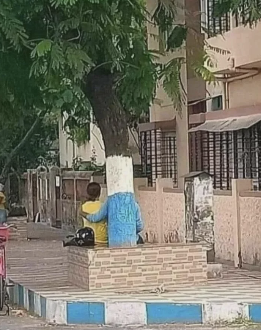 Two individuals sitting on a curb under a tree, one wearing a yellow top and the other in blue, with a backpack beside them