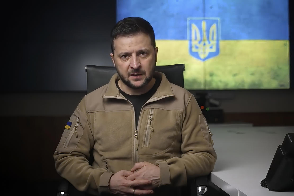 Ukrainian President Volodymyr Zelensky, seated in front of a blue and gold banner and wearing a beige zippered jacket, looks seriously into the camera..