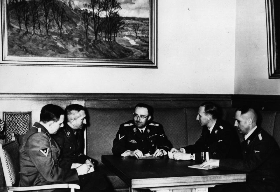 five men sit around a table and talk, they wear uniforms, a large painting hangs on the wall in the background