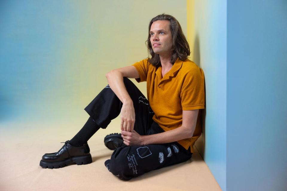 Aaron Jackson wears an orange shirt and dark pants while sitting on the floor next to a blue wall.