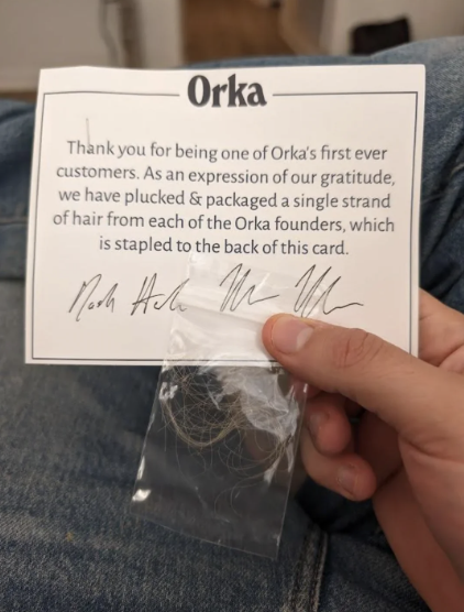 A hand holds a card and bag from Orka with a strand of hair, thanking customers for their support, with two signatures