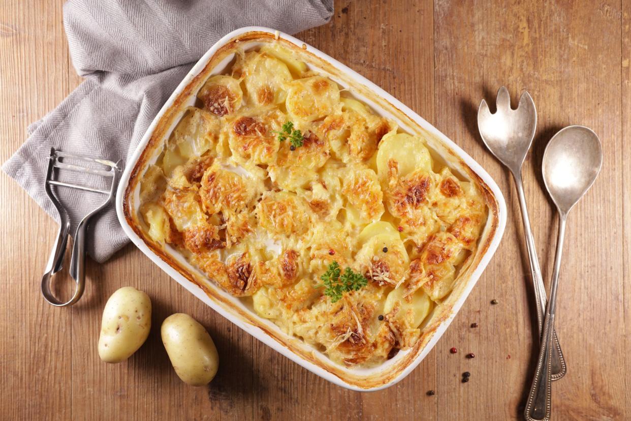 Potatoes au gratin in a white stoneware pan with cooking utensils and two potatoes on a rustic wooden table