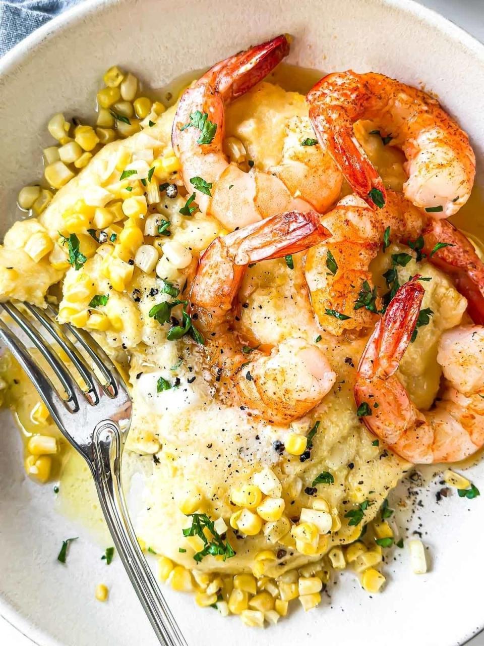 A bowl of creamy polenta topped with shrimp, corn, herbs, black pepper, and a fork on the side