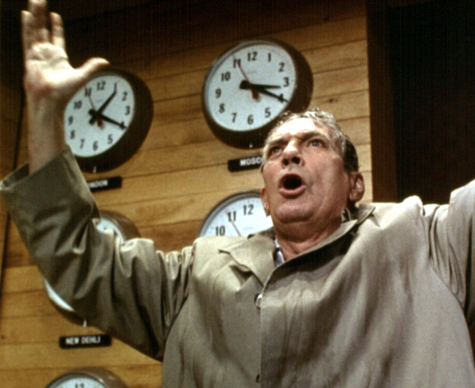 Peter Finch shouts in front of a wall of clocks drenched in a trench coat