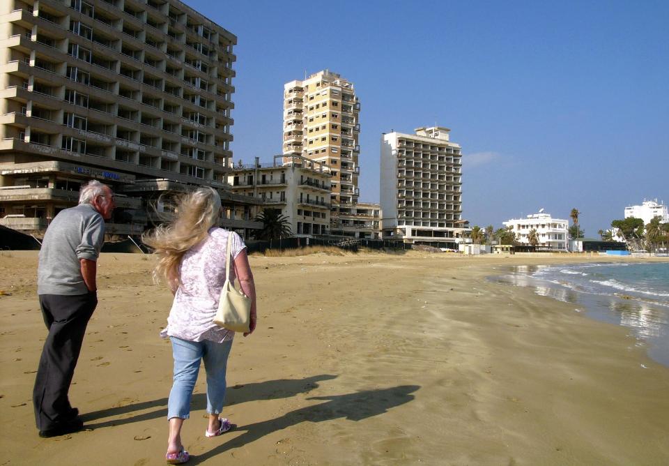 A couple walk on the beach by the deserted hotels in an area used by the Turkish military in the Turkish occupied area in abandoned coastal city of Varosha, in Famagusta, in southeast of island of Cyprus, Friday, Jan. 17, 2014. Time virtually stopped in 1974 for the Mediterranean tourist playground of Varosha. When Turkey invaded Cyprus in the wake of a coup by supporters of union with Greece, thousands of residents fled, and chain-link fences enclosed a glamorous resort that it's said once played host to Hollywood royalty like Elizabeth Taylor. The town's crumbling, war-scarred beachfront hotels have become an emblem of the country's division between Turks and Greeks. In 40 years, few have set foot inside the town, which remains heavily guarded by the Turkish army and twists of barbed wire. But that grim scene could present a rare opportunity. Massachusetts Institute of Technology architecture professor Jan Wampler calls it the greatest challenge of his career: he and a team of architects, urban planners, business leaders and peace activists hope to rebuild an entire town to correct past errors and mold a sustainable, ecological habitat. (AP Photo/Petros Karadjias)