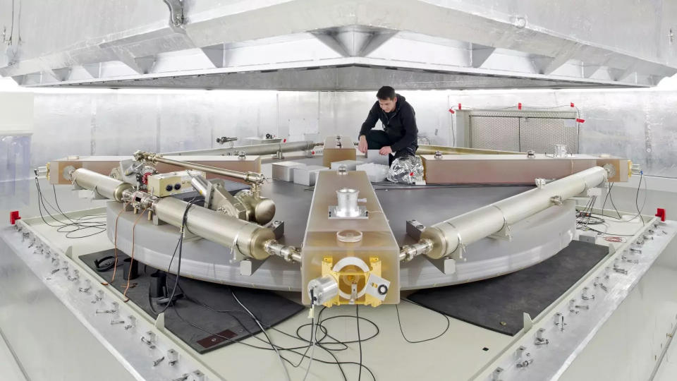 a man in black clothing sits atop a large, square metallic structure in a white-walled laboratory