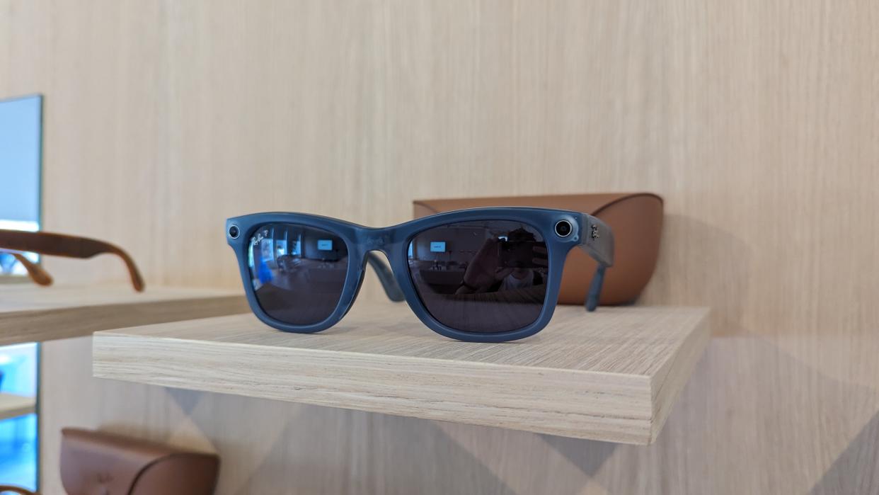  A blue pair of the Ray-Ban Meta Smart Glasses Collection on a wooden table in front of their charging case. 
