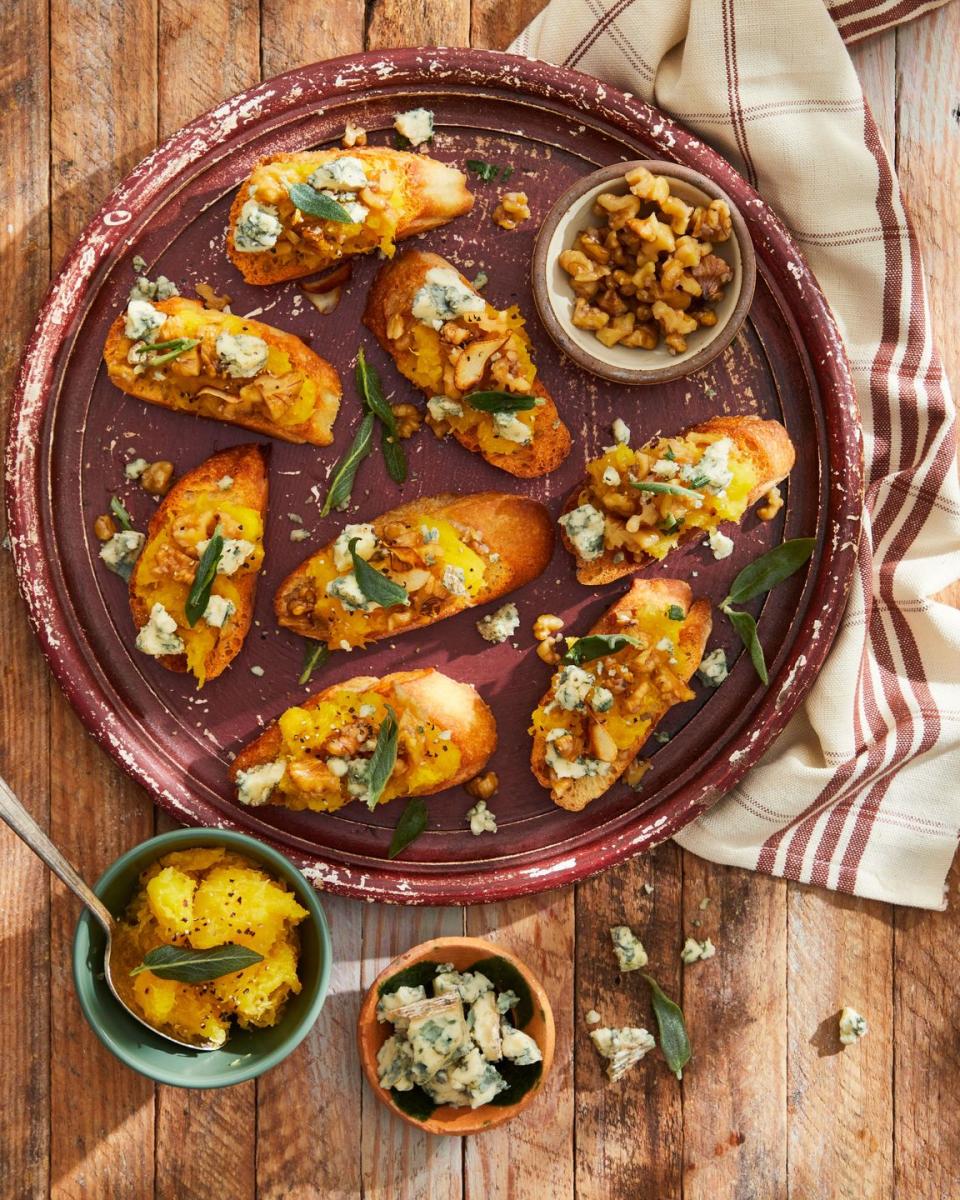 smashed acorn squash and garlicky walnut crostini on a dark red plate with small bowls of toppings on the side