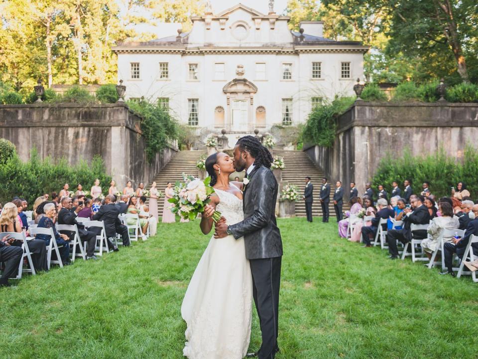 A bride and groom kiss at the end of their wedding altar in front of a mansion.