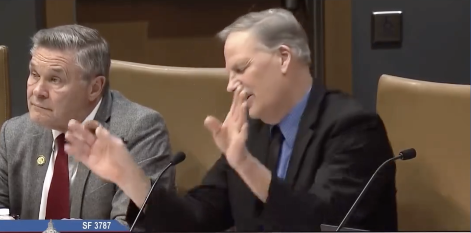 Two individuals seated behind microphones in a hearing, one is clapping and appears animated