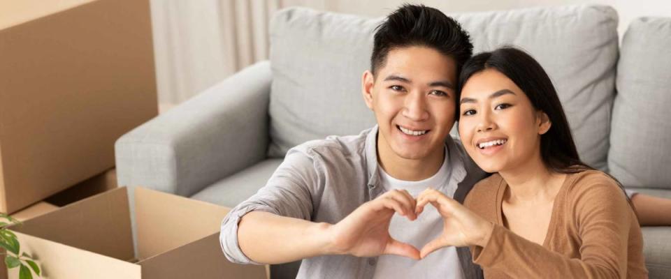 Love, Family, Home Concept. Portrait of cute asian couple sitting among moving boxes and making heart sign with fingers