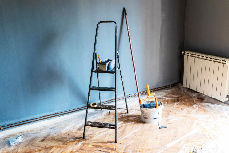 Painting tools and a step ladder sitting in an empty room prepared for renovation with a plastic drop cloth covering the hardwood floor. 