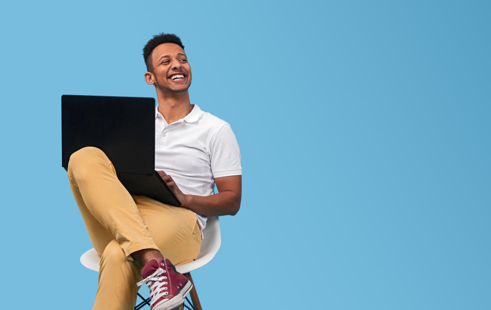 Young man smiling and looking at empty space while sitting on chair and using laptop against blue background