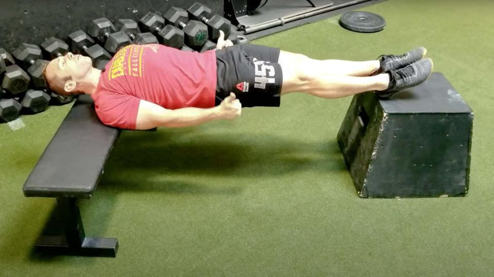 YouTube: Man performing a Chinese plank with upper back supported on a bench and heels supported on box during gym workout