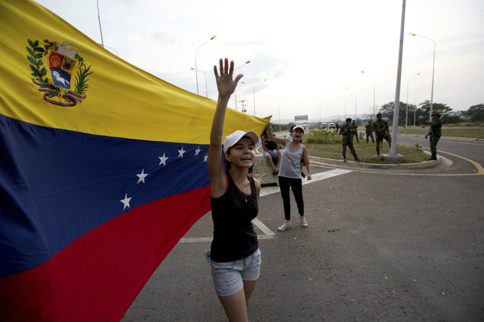 Woman shouts "Welcome humanitarian aid" as he waives Venezuelan flag in front aof a group of Venezuelan Army soldiers and National Guard officers blocking the main access to the Tienditas International Bridge that links Colombia and Venezuela, near Urena, Venezuela, Thursday, Feb. 7, 2019. Venezuela's opposition leaders requested the shipments and vowed to bring them inside the troubled nation despite objections from embattled President Nicolas Maduro. (AP Photo/Fernando Llano)
