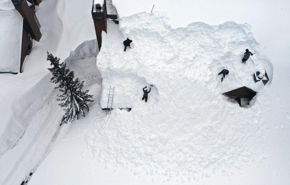 Small figures in dark clothing are seen in a broad field of white snow, along with the windows of a building's upper story.