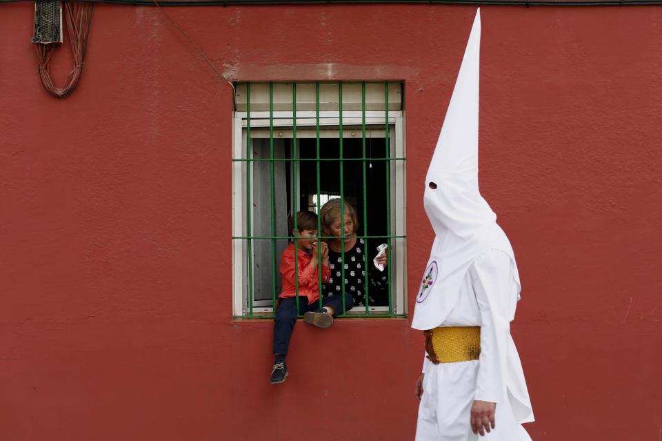 A woman and a boy look from a window as a penitent of San Gonzalo brotherhood walks past them during Holy Week in the Andalusian capital of Seville