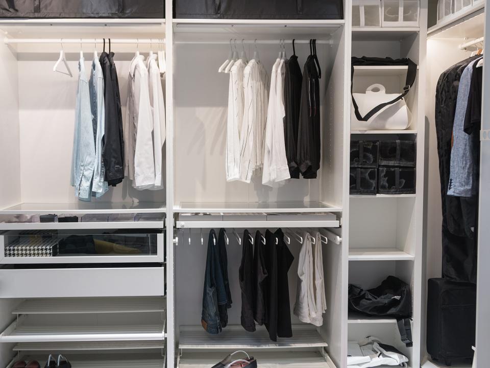A closet with white shelves holds a capsule wardrobe with white, black, gray, and chambray clothing items and accessories.