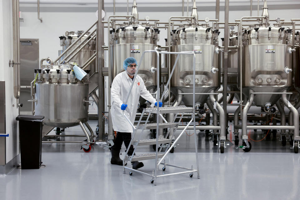 A technician in a white coat, blue nitrile gloves and a blue hair net pushes a trolley past an array of gleaming vats.