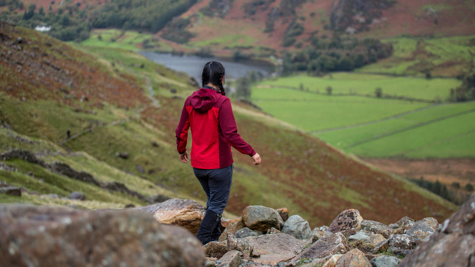 A woman wearing a Patagonia Women’s R1 CrossStrata Hoody is walking away from the camera, across a rocky hillside.